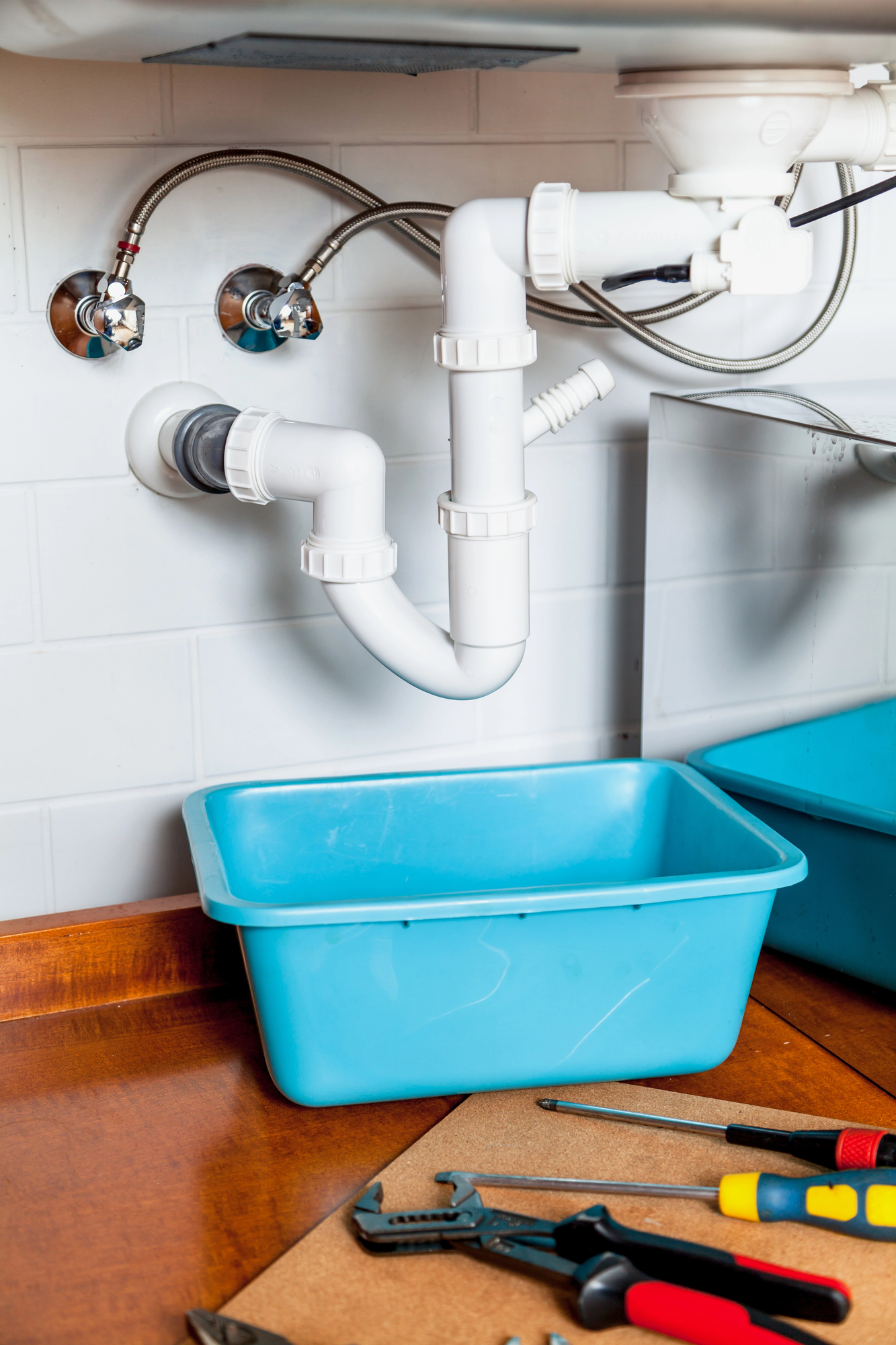 Plumbing pipes under a sink with a blue plastic container placed below, surrounded by various hand tools on a wooden surface.