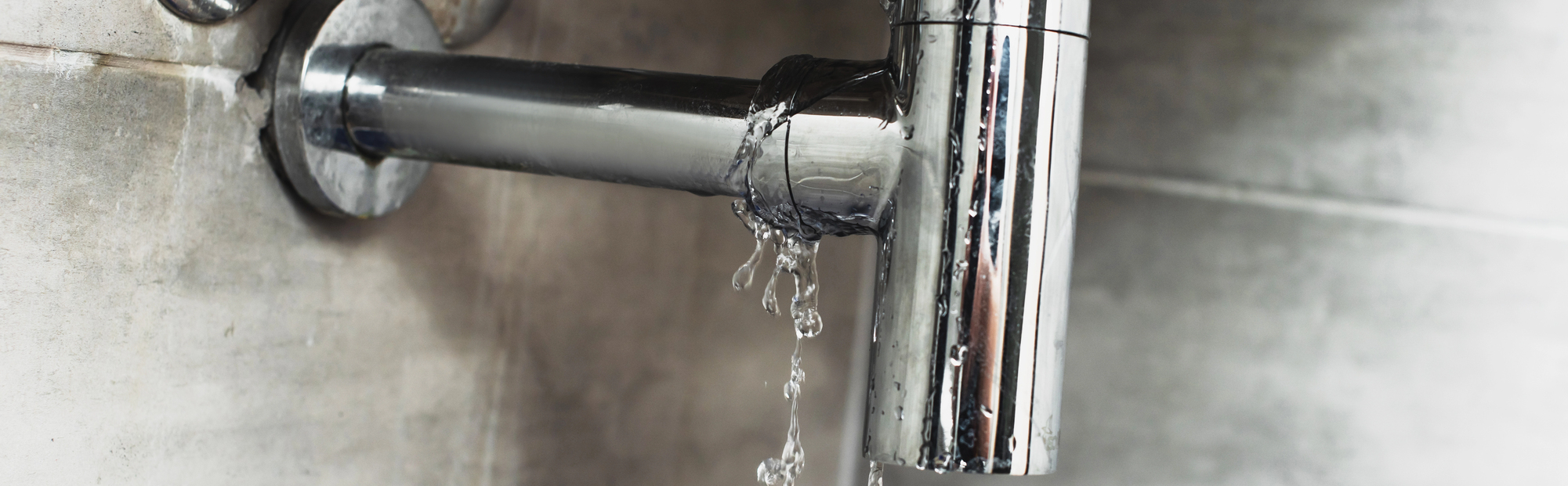 A chrome faucet mounted on a tiled wall leaking water, with several droplets falling from the spout.