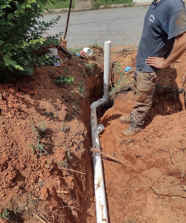 A person stands next to a partially installed PVC pipe system in a dirt trench, with tools and additional pipe sections nearby.