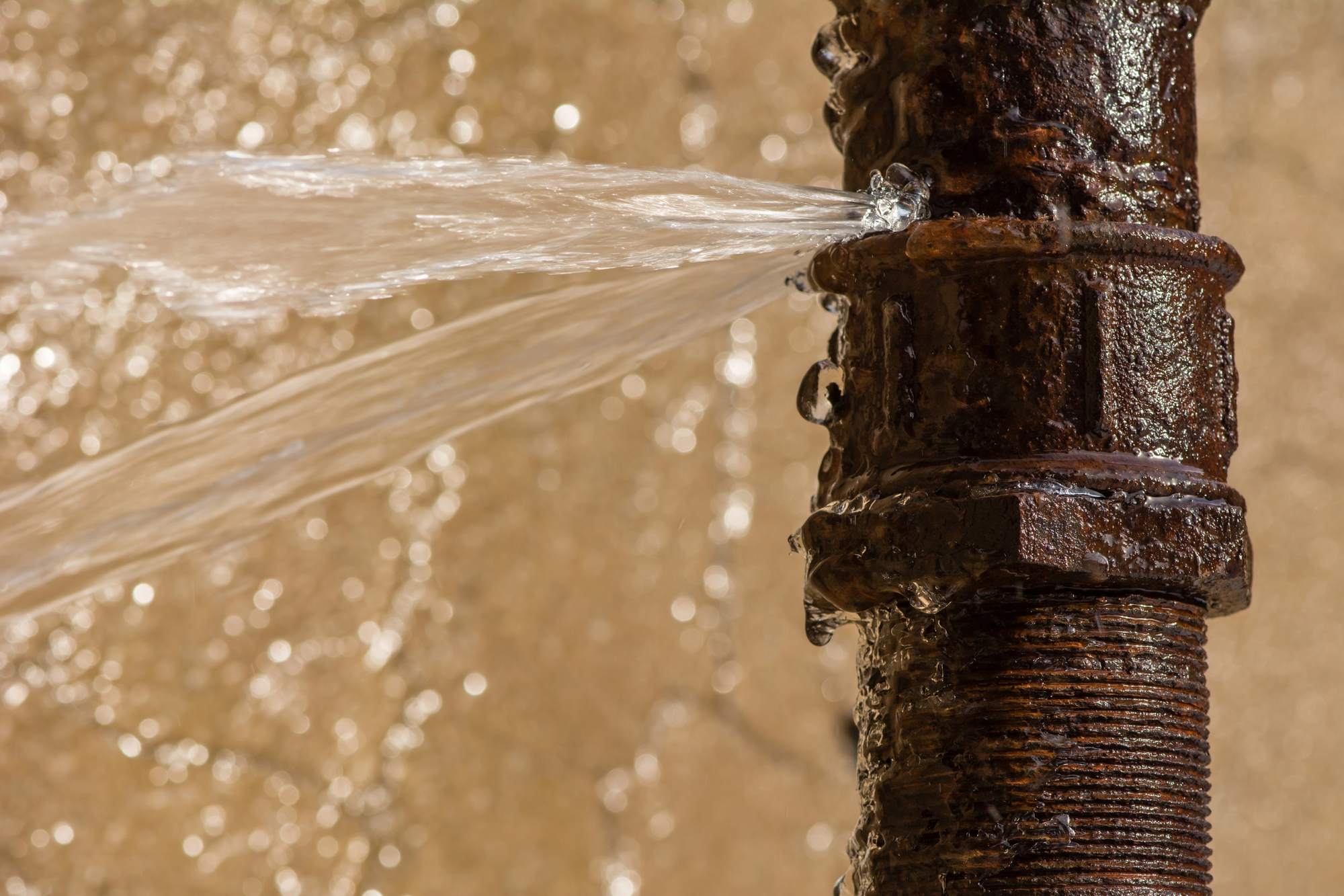 Water spraying from a crack in a rusty metal pipe, indicating a leak.
