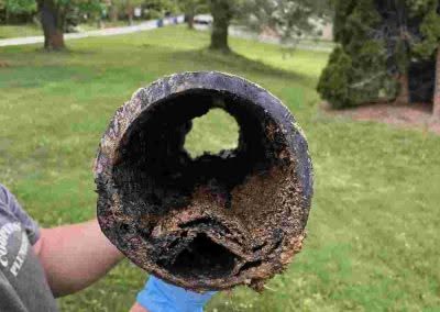 A person holds a corroded and rusty metal pipe with significant buildup and damage, viewed from one open end, outdoors on grass.