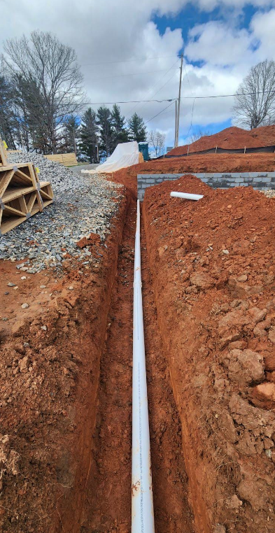 White PVC pipe is laid in a trench through red soil at a construction site, with gravel and stacked materials visible on the left and a partially built wall in the background.