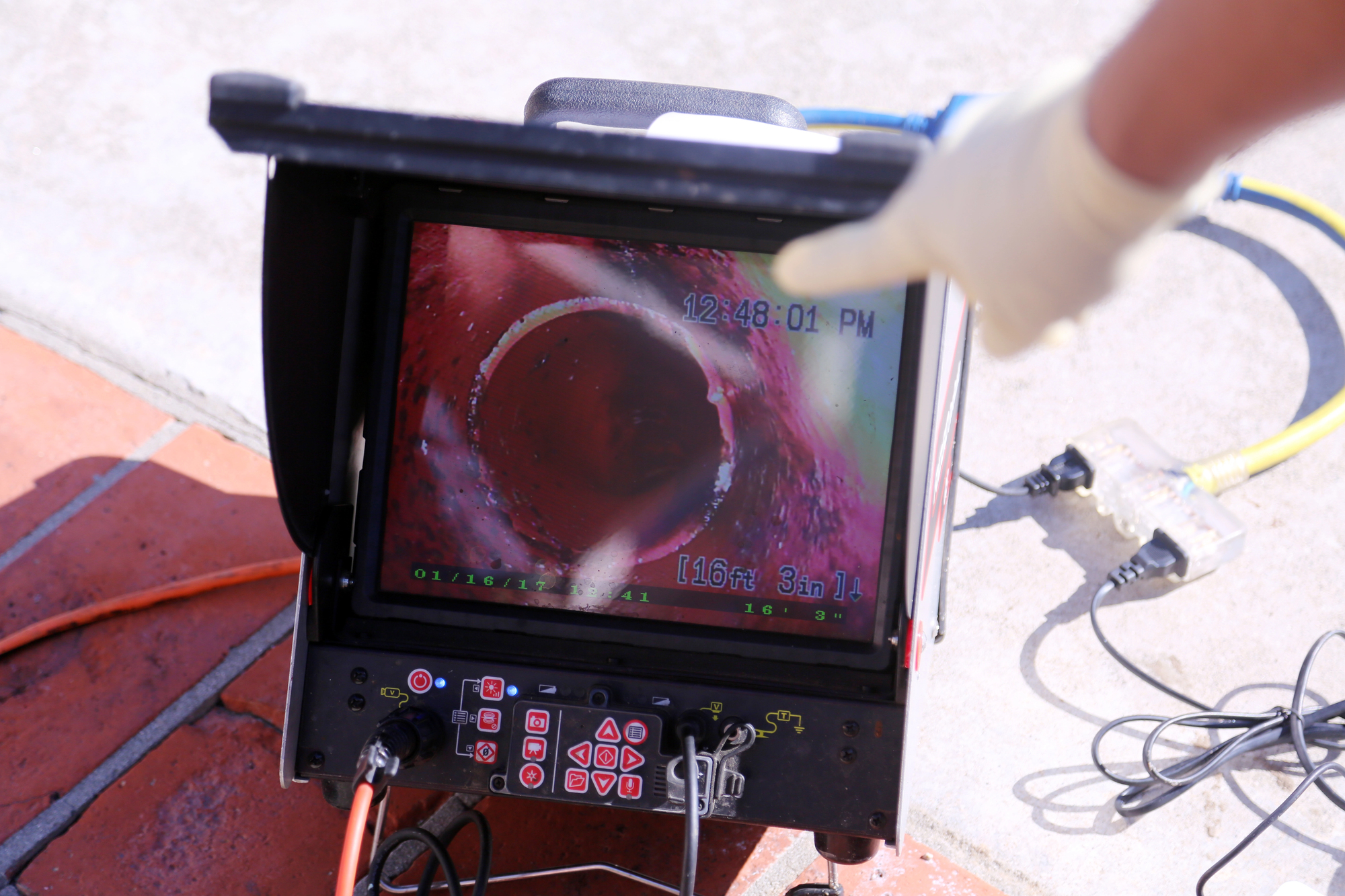 A gloved hand points at a monitor displaying a live feed from a pipe inspection camera, showing the inside of a pipe.