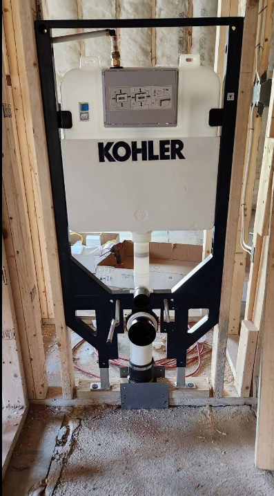 Kohler in-wall toilet tank and carrier system installed in wooden wall framing during construction, with visible piping and insulation.